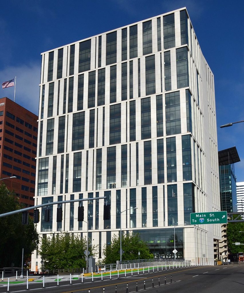 A picture of a recently constructed multiple story building with six segments of multiple floors each with its own series of alternating vertical stripes of windows and white wall of varying widths. The stripes of each segment do not align with the stripes of the segment below or above. The sky behind the building is blue and other buildings can be seen to the right and left. In the lower right corner is an asphalt street with a green sign with white text reading "Main St. To I-5 South."