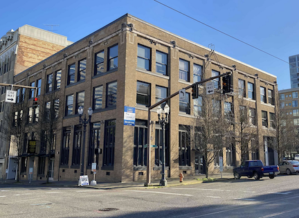 A brown brick building on the corner of two intersecting streets with a 'For Lease' sign hanging on one side. Vehicles are parked along one side of the building.